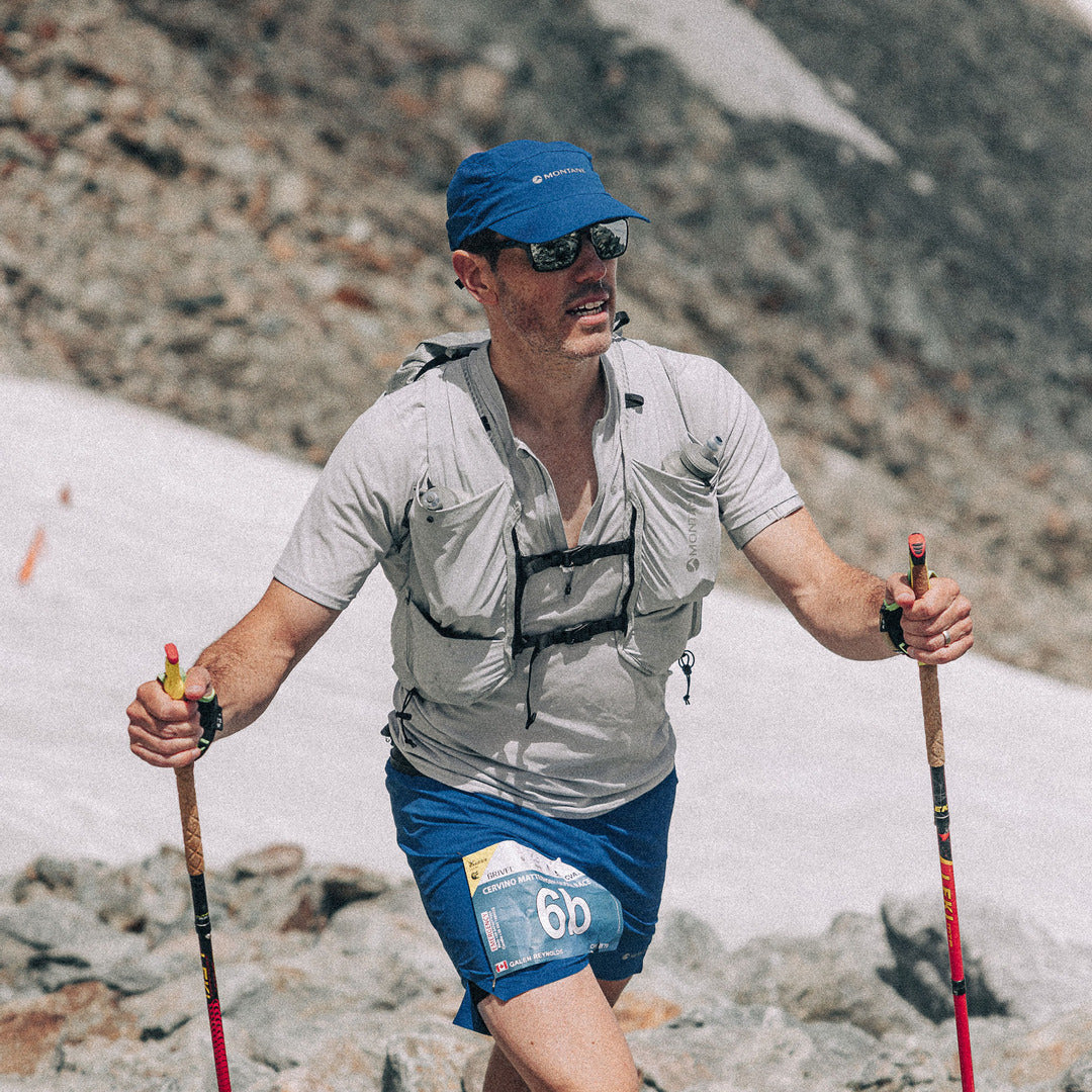 A male trail runner wearing a light gray compression vest, blue shorts, a blue cap, and sunglasses, holding trekking poles while running through rocky, snowy mountain terrain.