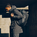 Person wearing a dark coat and carrying a backpack against a dark background