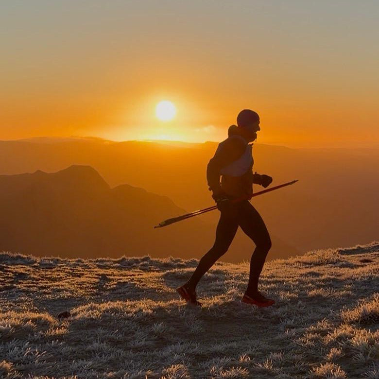 A runner carrying poles is silhouetted against a dramatic orange sunset over a mountain range, running across a frosty, grassy ridge.